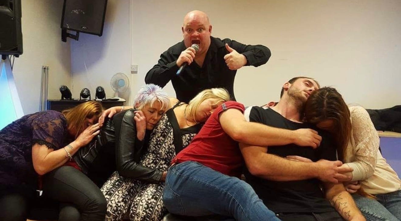 Jason on the microphone giving a thumbs up behind a row of hypnosis volunteers asleep on each other’s shoulders