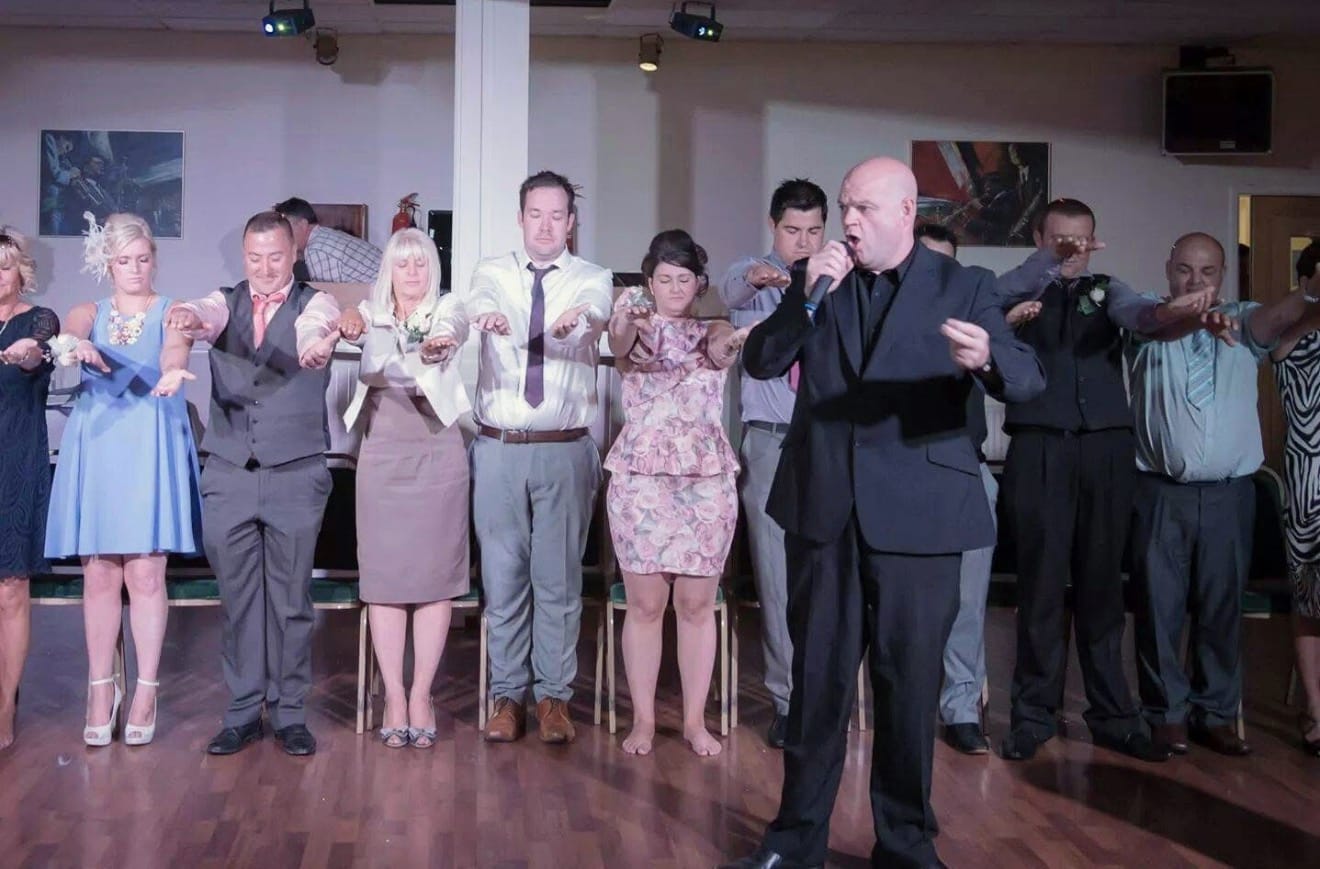 Jason on the microphone directing a line of wedding guests who are holding their arms out during a comedy hypnosis show