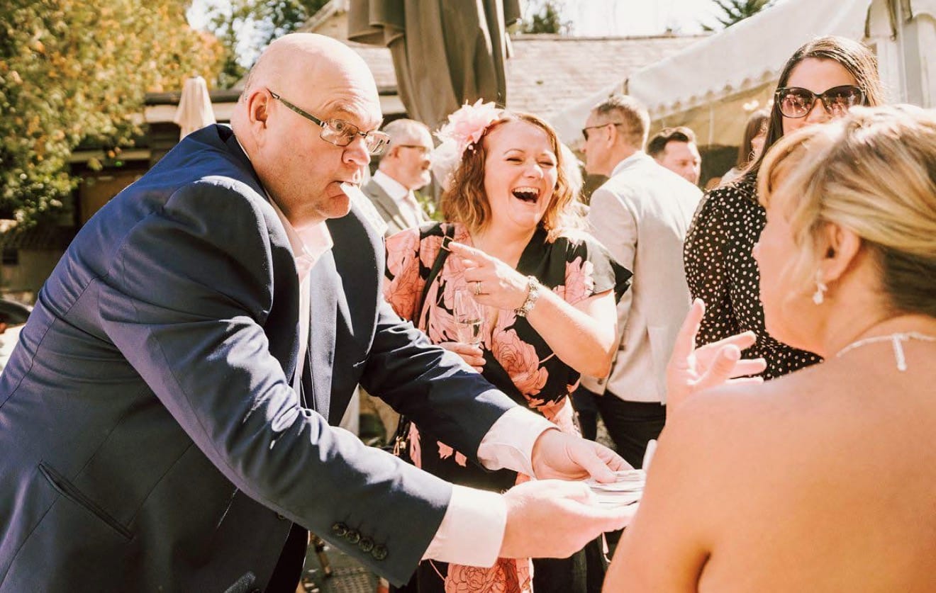 Jason performing card magic for wedding guests at an outdoor drinks reception while one guest laughs